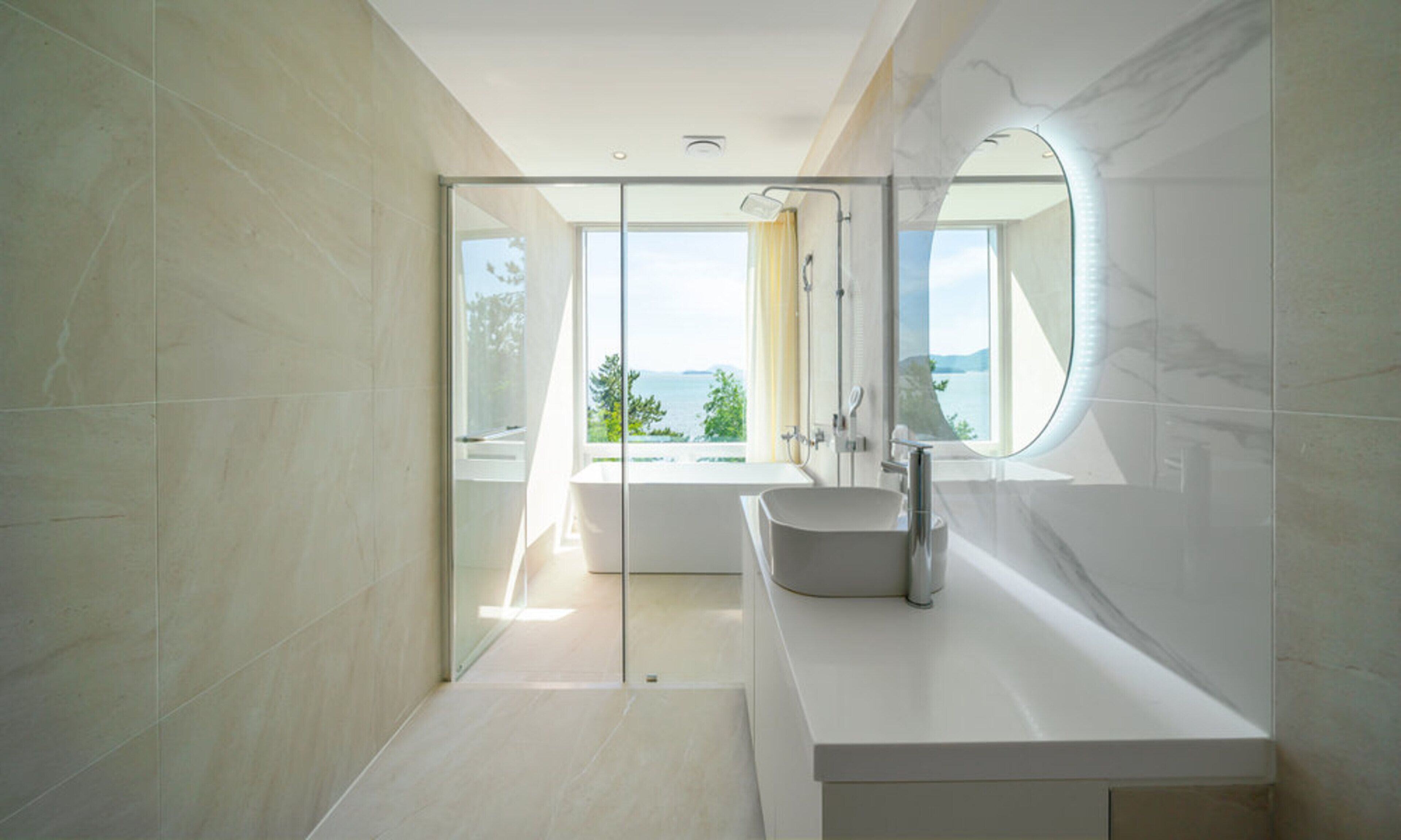 A modern bathroom with a large window, a round mirror, and a white sink.