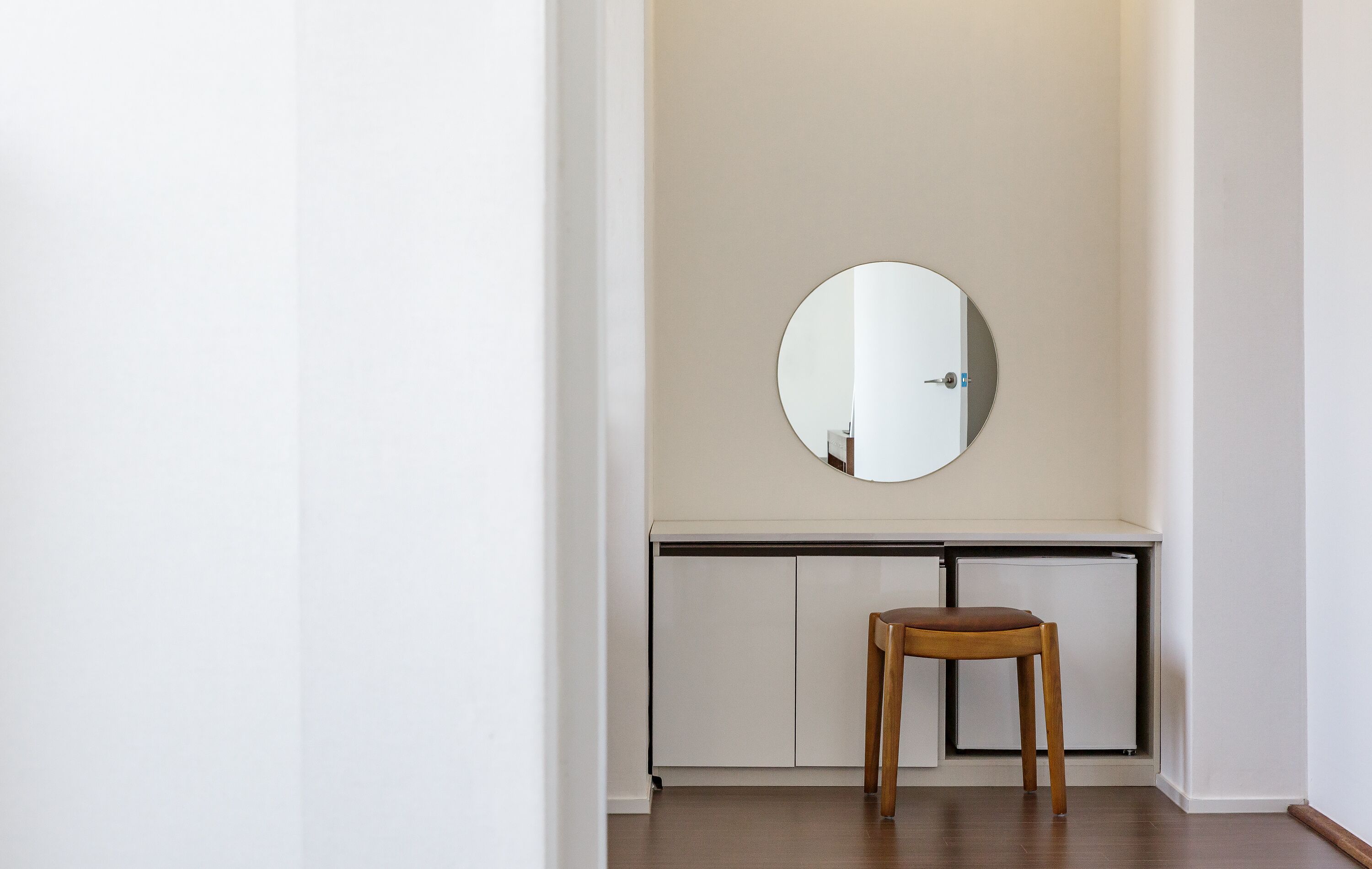 A minimalist room with a round mirror on the wall, a wooden chair, and a white cabinet below.