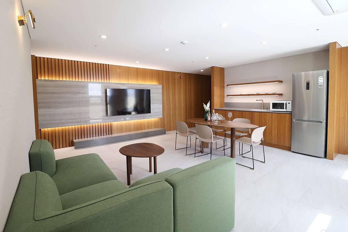 Ein modernes Wohnzimmer mit Flachbildfernseher, grünen Sofas, einer Essecke mit Tisch und Stühlen sowie einer Küche mit Kühlschrank und Mikrowelle.