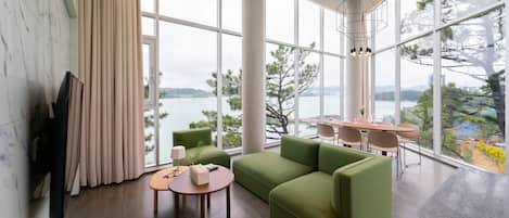 Een moderne woonkamer met een groene bank, een houten salontafel, en een eethoek met tafel en stoelen. Grote ramen bieden uitzicht op bomen en een waterpartij.