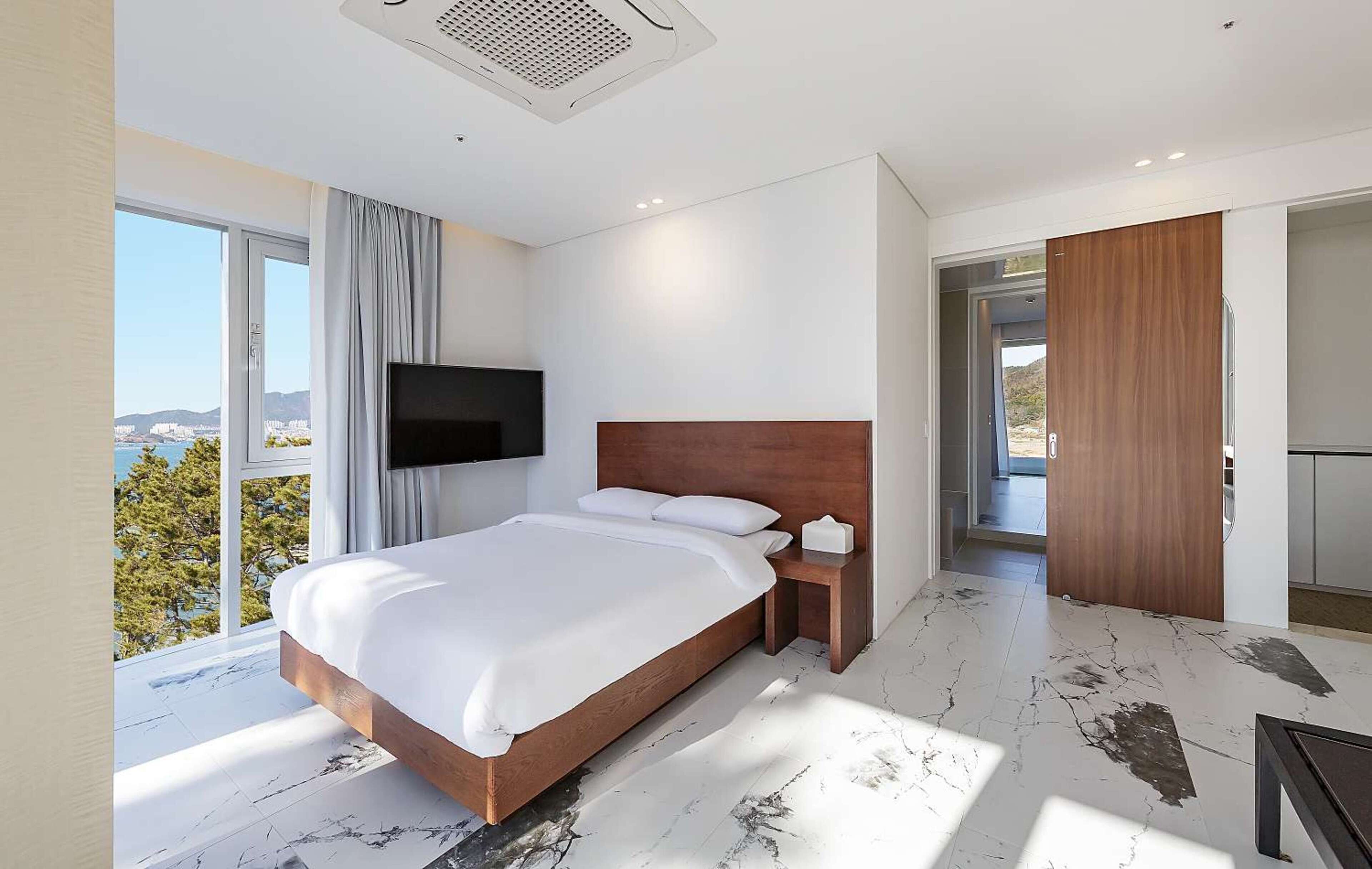 A modern hotel room with a large bed, a flat-screen TV, a bedside table, and a view of the outdoors.