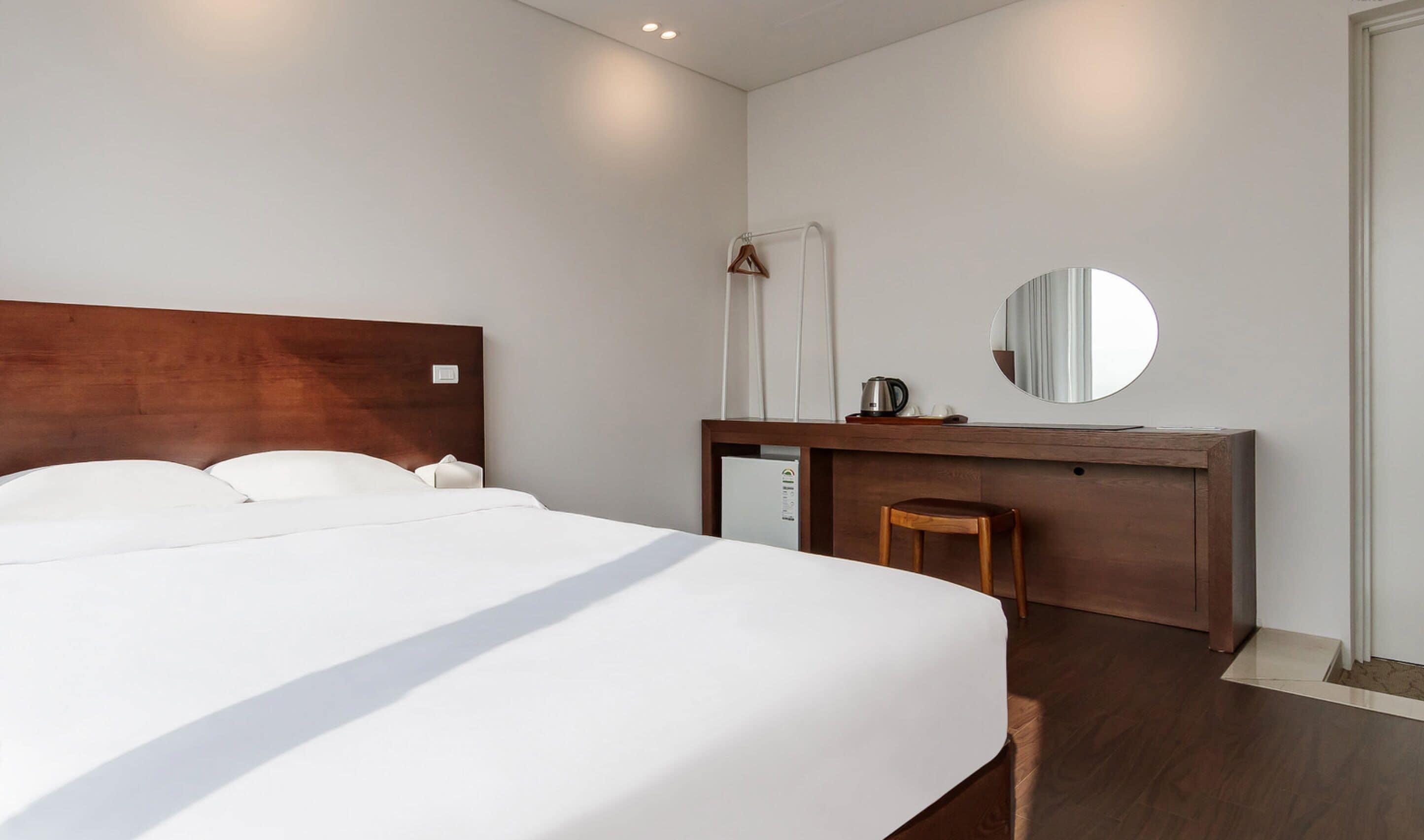 A modern hotel room with a large bed, a wooden desk, a chair, a mirror, and a small kitchenette.