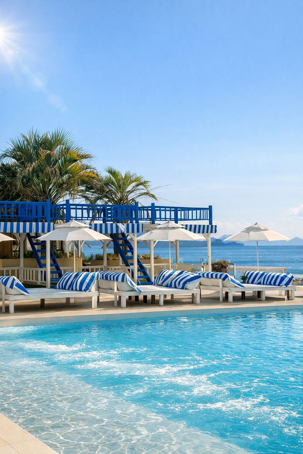 A poolside area with lounge chairs and umbrellas, a covered seating area, and a view of the ocean.