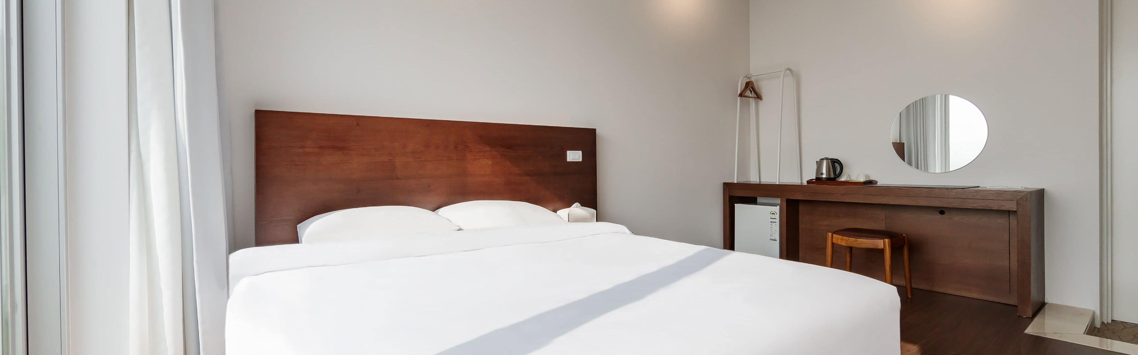 Une chambre d’hôtel moderne avec un grand lit, une tête de lit en bois, un bureau avec un miroir et un petit tabouret.