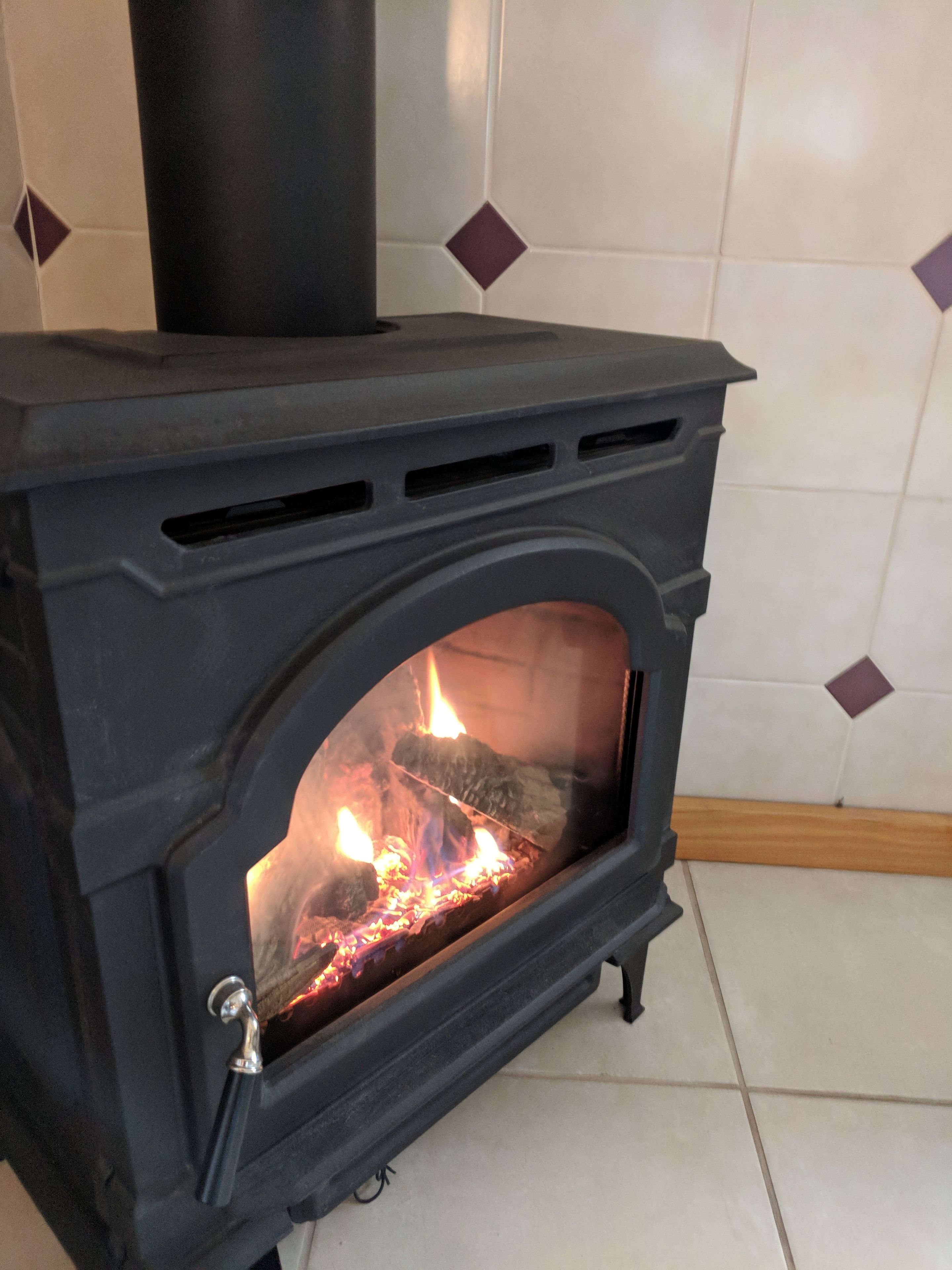 A black wood-burning stove with a glass door, set against a tiled wall with diamond patterns.