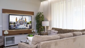 A modern living room with a flat-screen TV mounted on the wall, a beige sofa, a small side table with a lamp, and a potted plant.