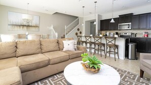 A modern living room with a beige sofa, a round coffee table with a plant, a dining area with a small island, and a kitchen with dark cabinets and stainless steel appliances.
