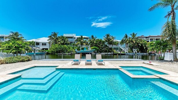 A pool area with clear blue water, lounge chairs, and a view of a marina with boats and buildings in the background.