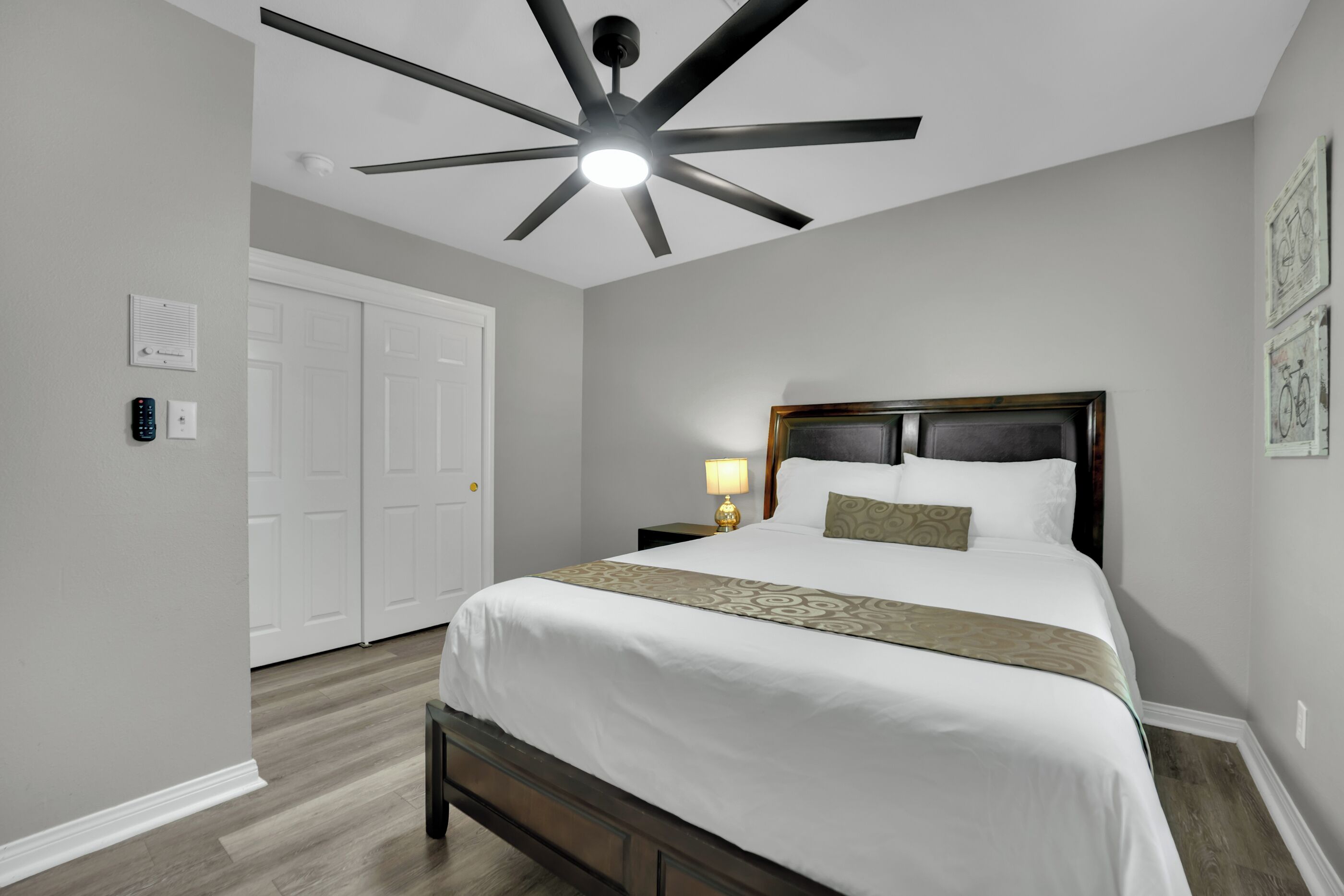 Un dormitorio con cama, ventilador de techo, mesita de noche, lámpara y puerta.