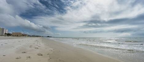 Une plage où l’on voit des empreintes de pas menant à l’eau, un ciel nuageux et des bâtiments en arrière-plan.