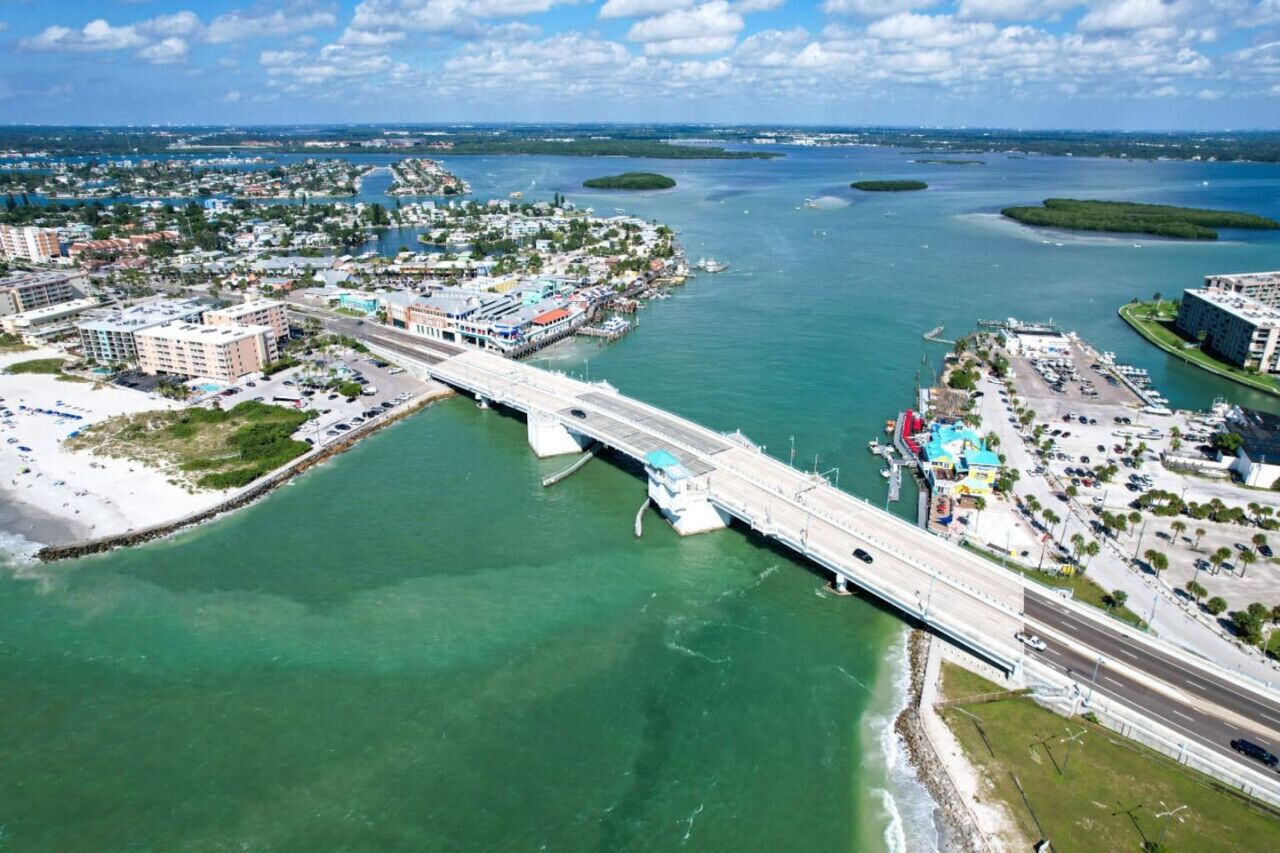An aerial view of a coastal city with a prominent bridge connecting two land masses, clear blue waters, and a mix of residential and commercial buildings.