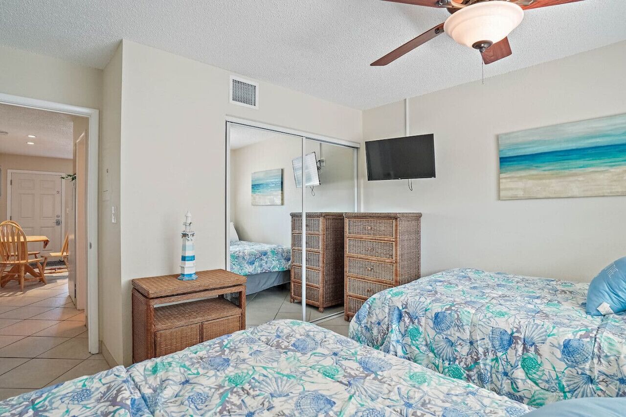 A bedroom with two beds, a ceiling fan, a TV, and a beach-themed painting.