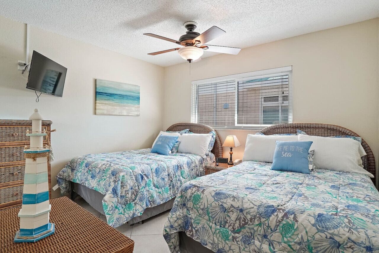 A bedroom with two beds, a ceiling fan, a TV, a window with blinds, and a decorative lighthouse model.
