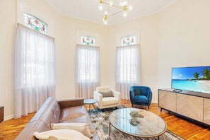 Een woonkamer met een leren bank, een witte fauteuil, een blauwe fauteuil, een glazen salontafel en een televisie op een houten kast.