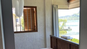 Une chambre avec un lit, une fenêtre avec des volets en bois et une vue sur une étendue d’eau et de la verdure.