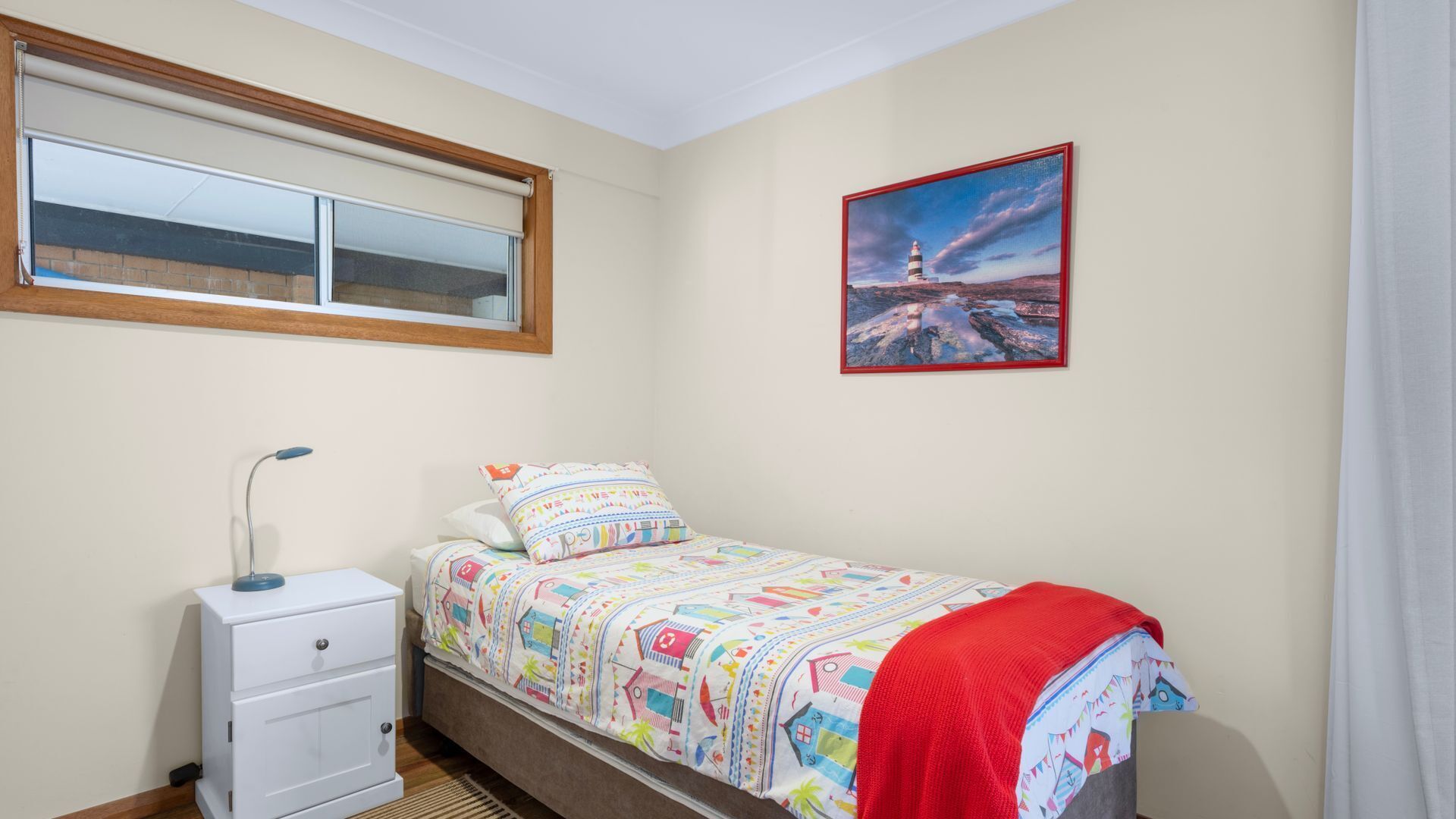 Een slaapkamer met een bed, een nachtkastje, een lamp, een raam met jaloezieën en een schilderij aan de muur.
