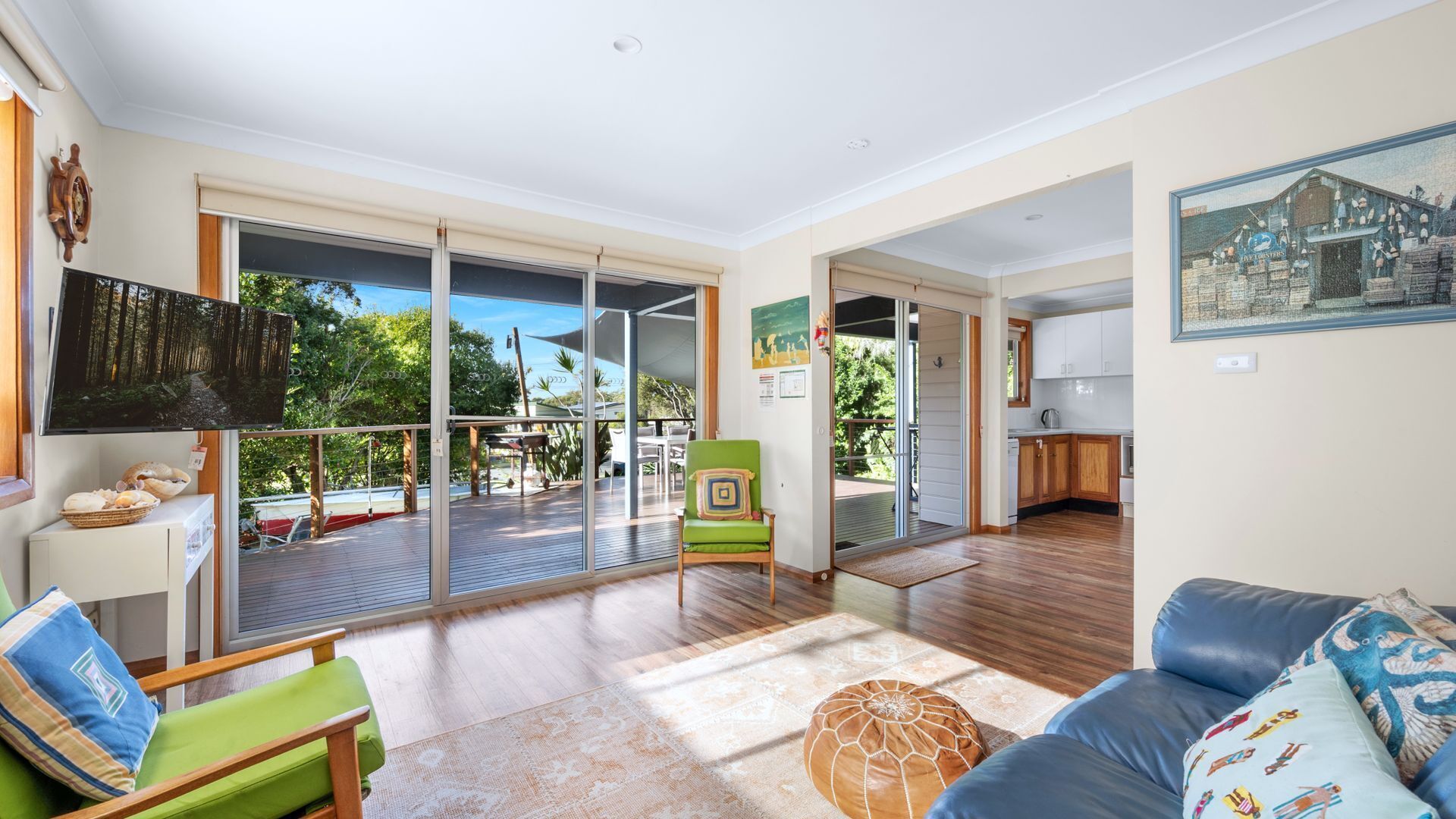 A spacious living room with a large sliding glass door leading to an outdoor deck, a cozy seating area with a green chair and ottoman, and a kitchen visible through an open doorway.