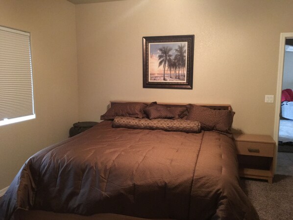A bedroom with a large bed, a nightstand, a drawer, a framed picture on the wall, and a window with blinds.