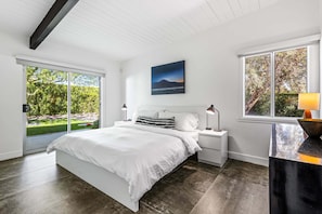 A bedroom with a large bed, a nightstand, a desk, a window with a view of greenery, and a painting on the wall.