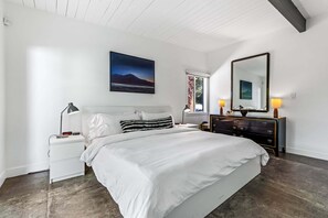 Un dormitorio con cama, mesitas de noche, cómoda, un espejo grande y un cuadro en la pared.