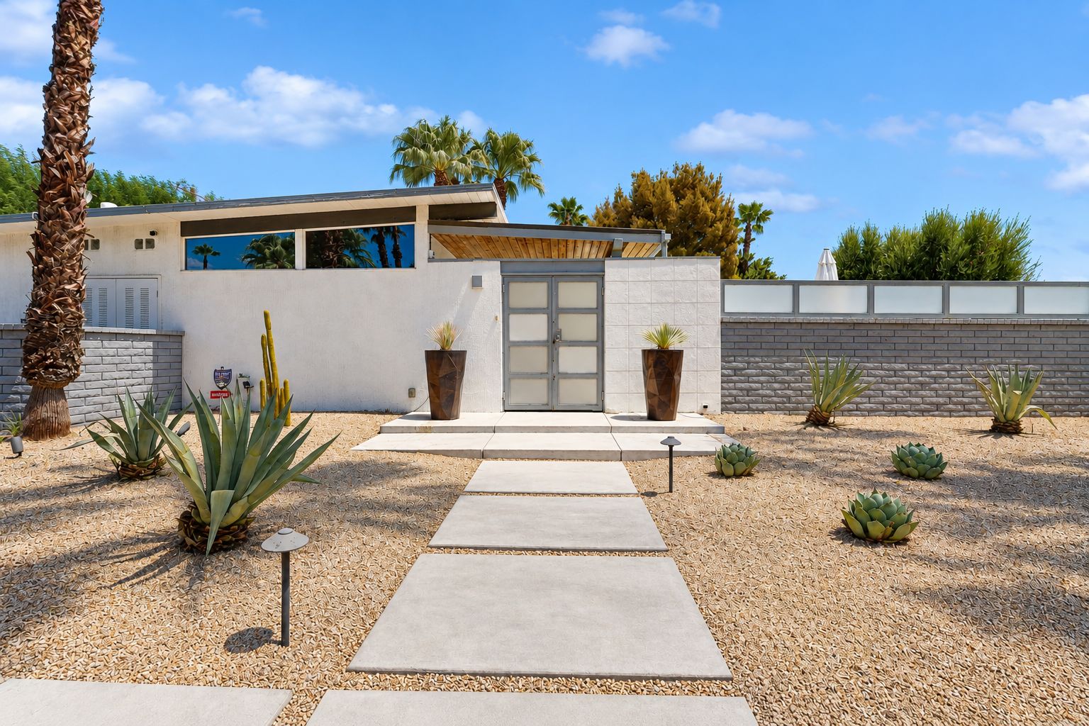 A modern house with a concrete pathway leading to a glass door entrance, surrounded by various plants and a stone wall.