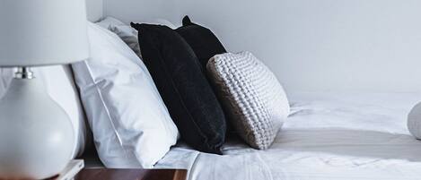 A neatly made bed with white bedding, a bedside lamp, and a stack of books on a wooden nightstand.