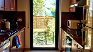 Una cocina con armarios de madera, un lavamanos de acero inoxidable, un microondas y un refrigerador. Hay una puerta que da a un balcón con vistas a zonas verdes.
