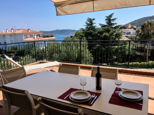 Une table à manger dressée avec des assiettes, des couverts et une bouteille de vin, offrant une vue panoramique sur une charmante ville côtière et la mer.