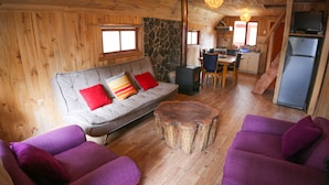 A cozy cabin interior with a sofa, two armchairs, a wooden coffee table, a stone wall, a kitchen area, and a large window.