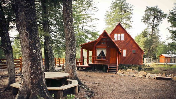 A rustic cabin with a wooden deck and a picnic table, surrounded by trees.