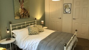 Un dormitorio con una cama blanca, dos almohadas, una mesita de noche con lámpara, un cuadro en la pared y dos puertas.