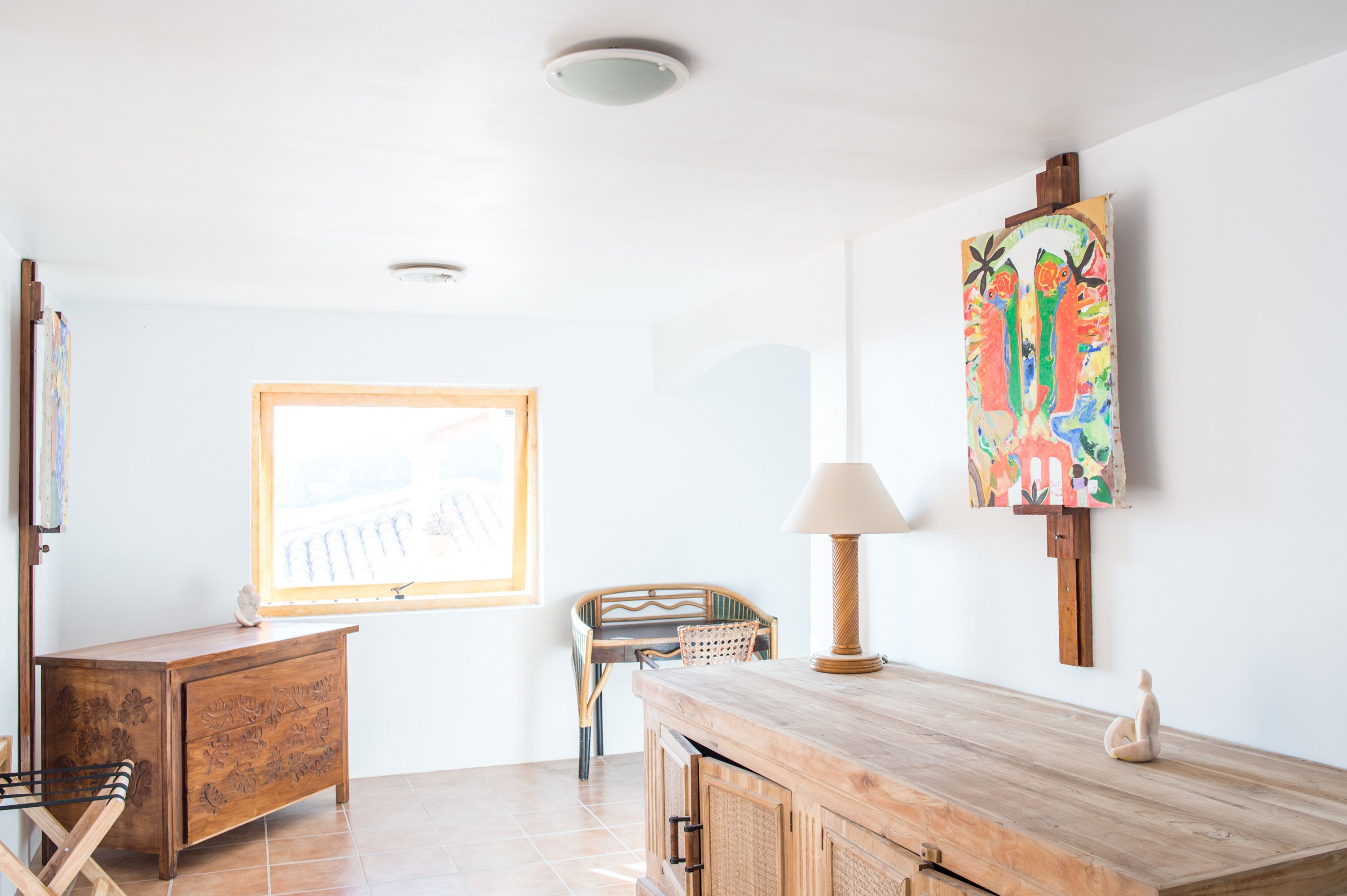 A room with a wooden desk, a chair, a lamp, a painting, and a window with a view.