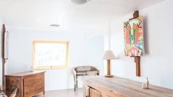 A room with a wooden desk, a chair, a lamp, a painting, and a window with a view.