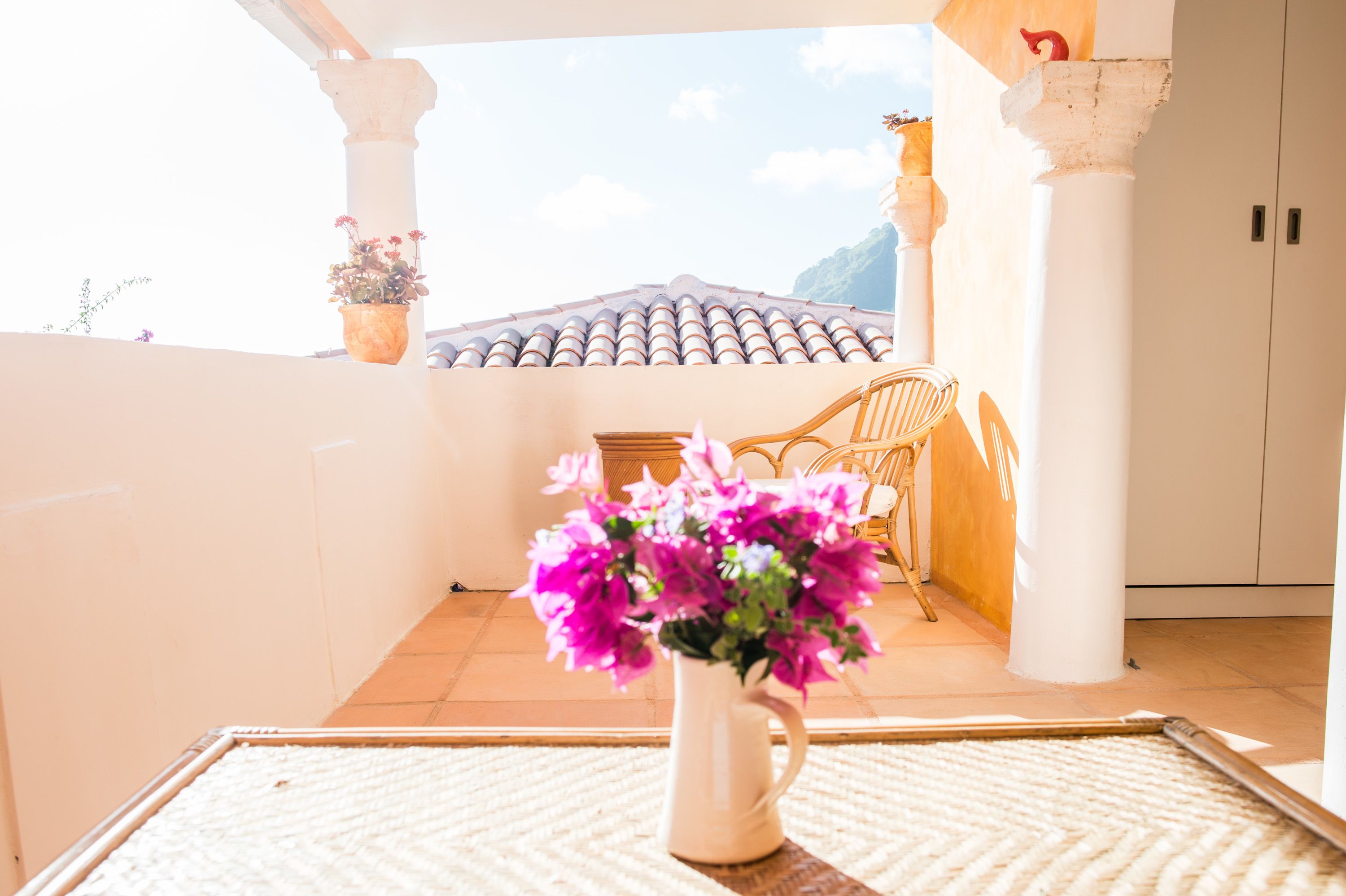 A balcony with a white vase of flowers, a wicker chair, and a tiled roof in the background.