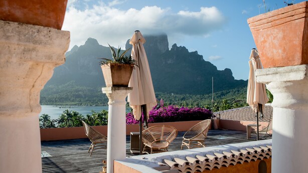 Una terraza con sillas de mimbre, plantas en macetas y sombrillas, con vistas a un paisaje montañoso y un cuerpo de agua.