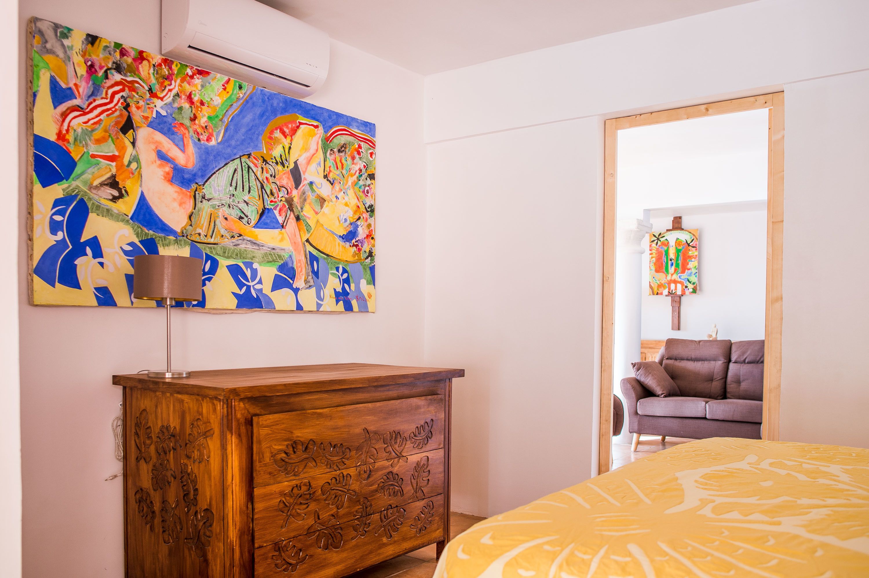 A bedroom with a wooden dresser, a bed with a yellow patterned bedspread, a colorful abstract painting on the wall, and a brown armchair in the background.