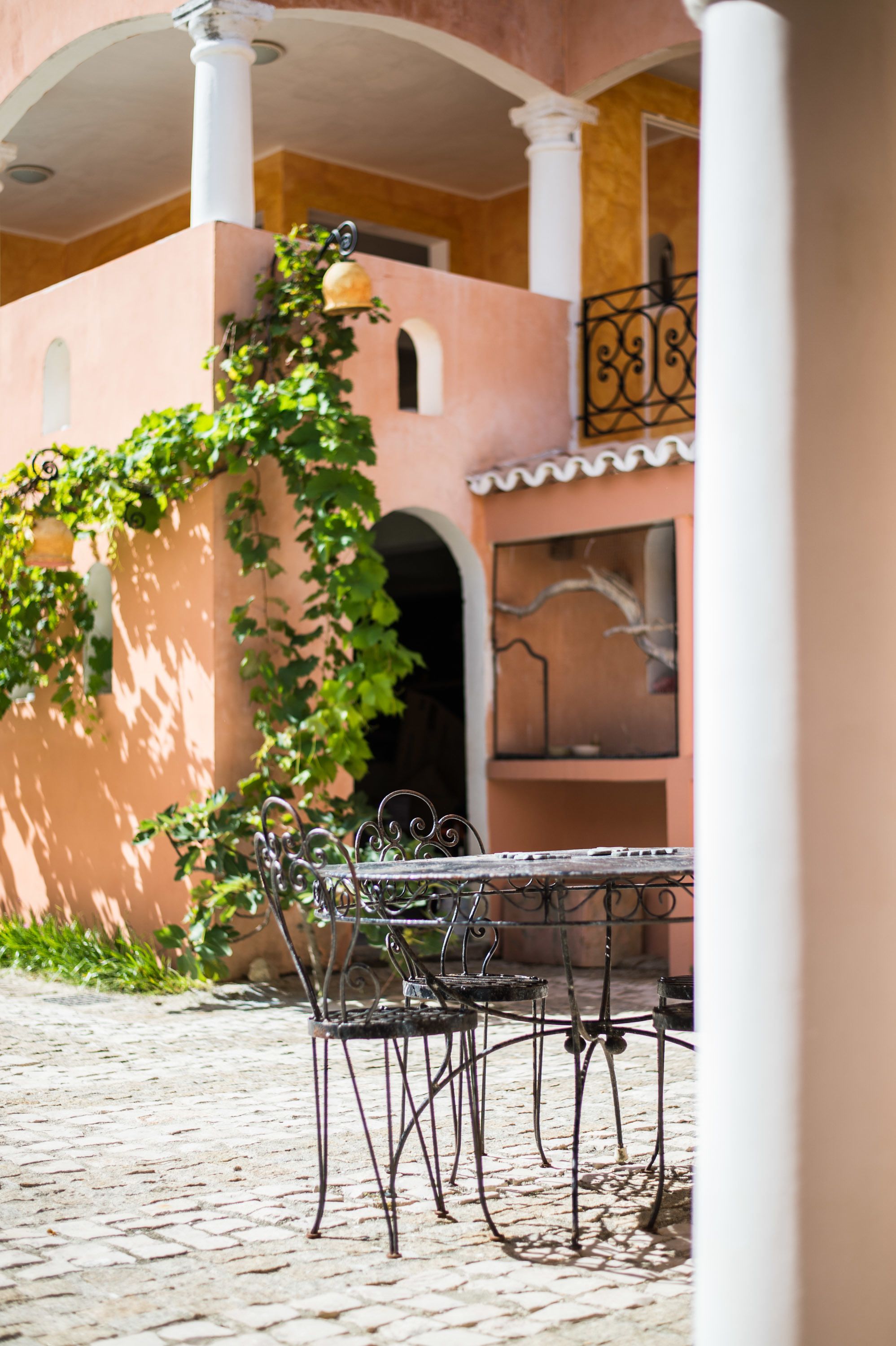 A cobblestone patio with a metal table and chairs, surrounded by a pink building with arches and a balcony.