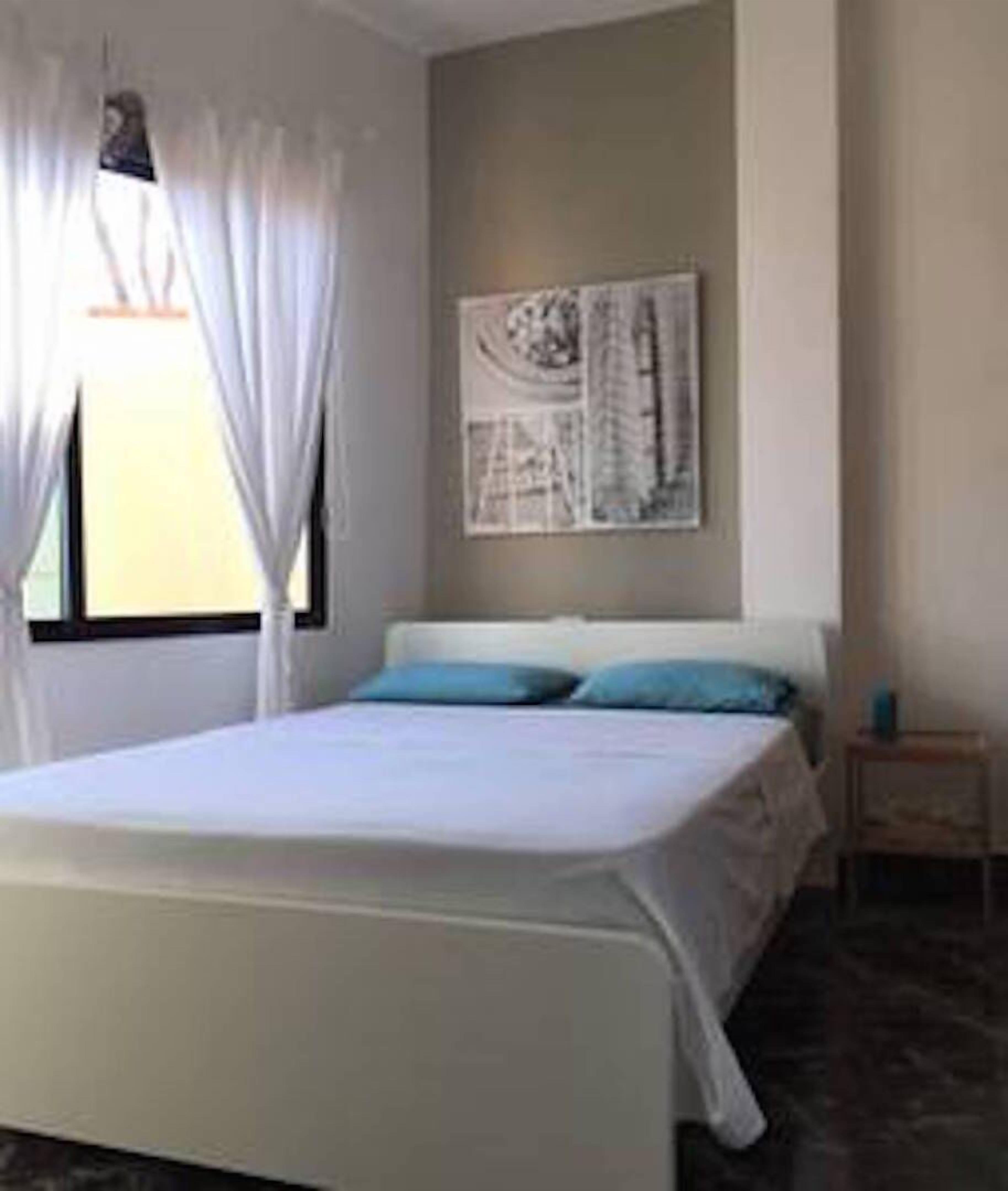 Une chambre à coucher avec un lit, des rideaux, une œuvre d’art accrochée au mur et une petite table de chevet.