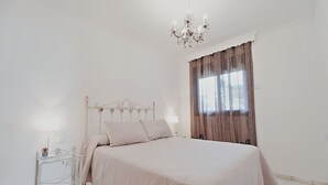 Een slaapkamer met een bed, nachtkastje, een kroonluchter en een raam met gordijnen.