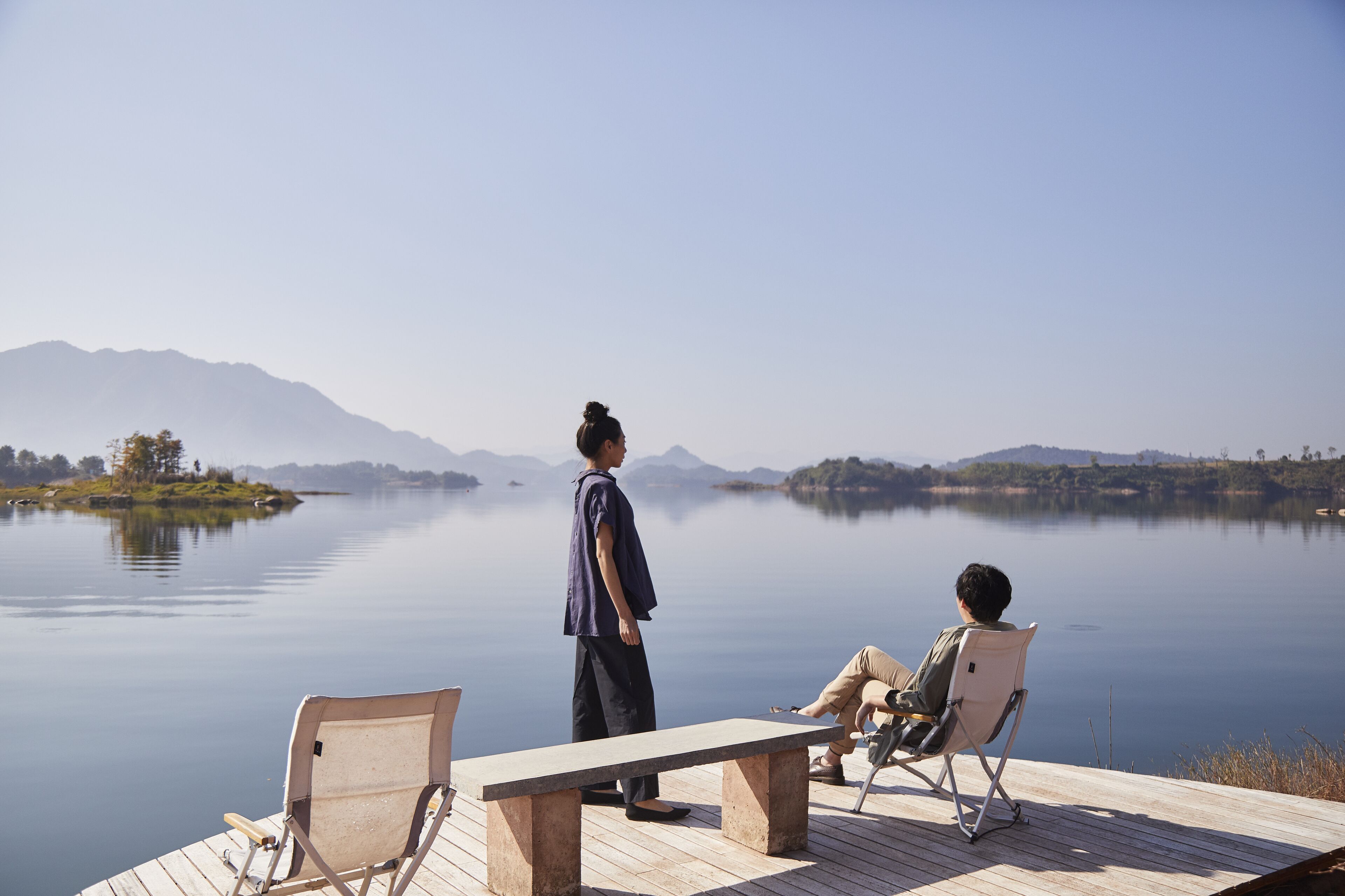 A wooden dock with two chairs and a bench overlooking a calm lake with mountains in the background.