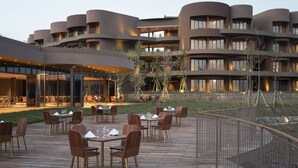 Un hotel moderno con fachada curva, zona de comedor al aire libre con mesas y sillas de madera, y un muro de contención de piedra.