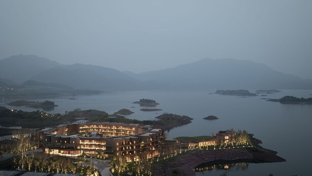 Un hotel moderno con un edificio central circular, rodeado por un área ajardinada con árboles y vistas a un lago tranquilo con varias pequeñas islas.