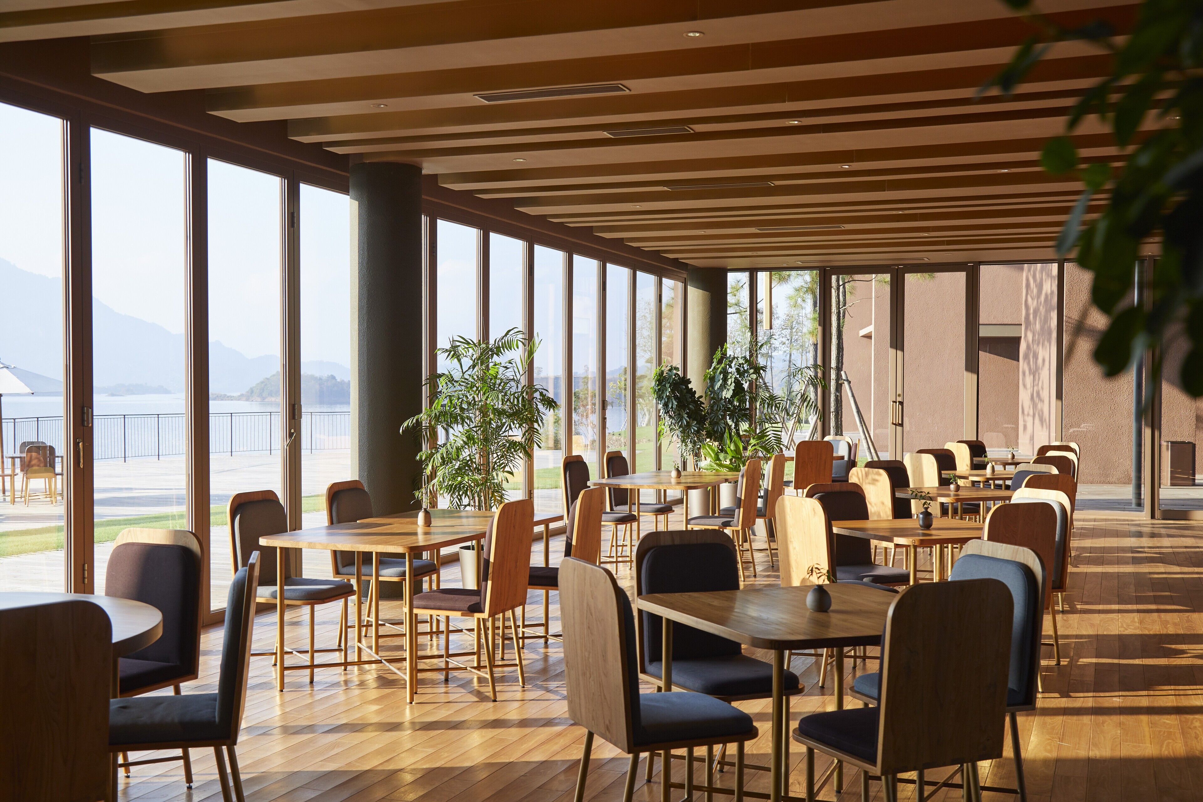 A spacious indoor area with large windows, wooden tables and chairs, and potted plants.