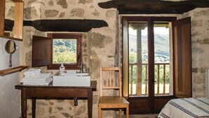 A rustic bathroom with a stone wall, wooden bench, and a window with a view of the countryside.