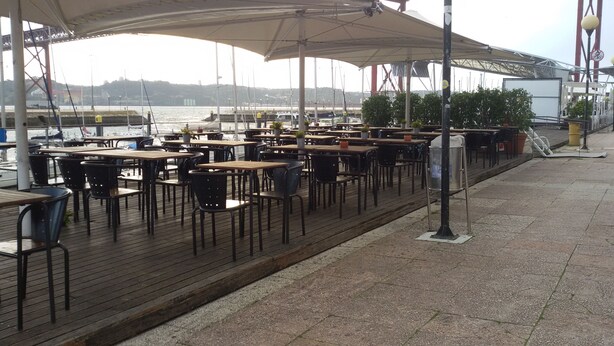 An outdoor café with wooden tables and chairs, located near a waterfront with a bridge in the background.