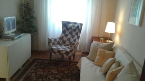 A living room with a patterned armchair, a sofa with cushions, a small table with a lamp, a television on a cabinet, and a plant.