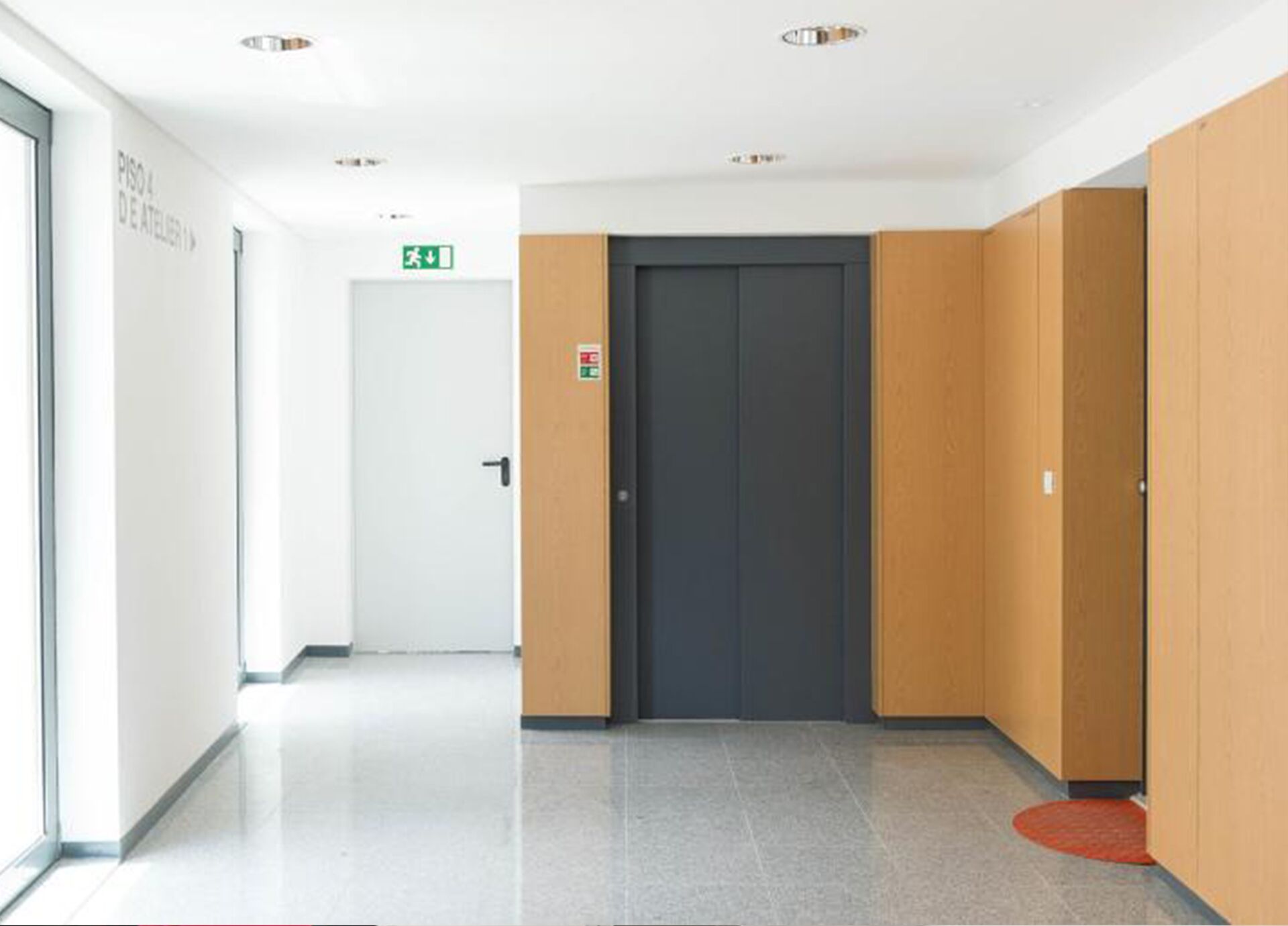 A modern hallway with a central elevator, a door with an emergency exit sign, and a glass wall on one side.