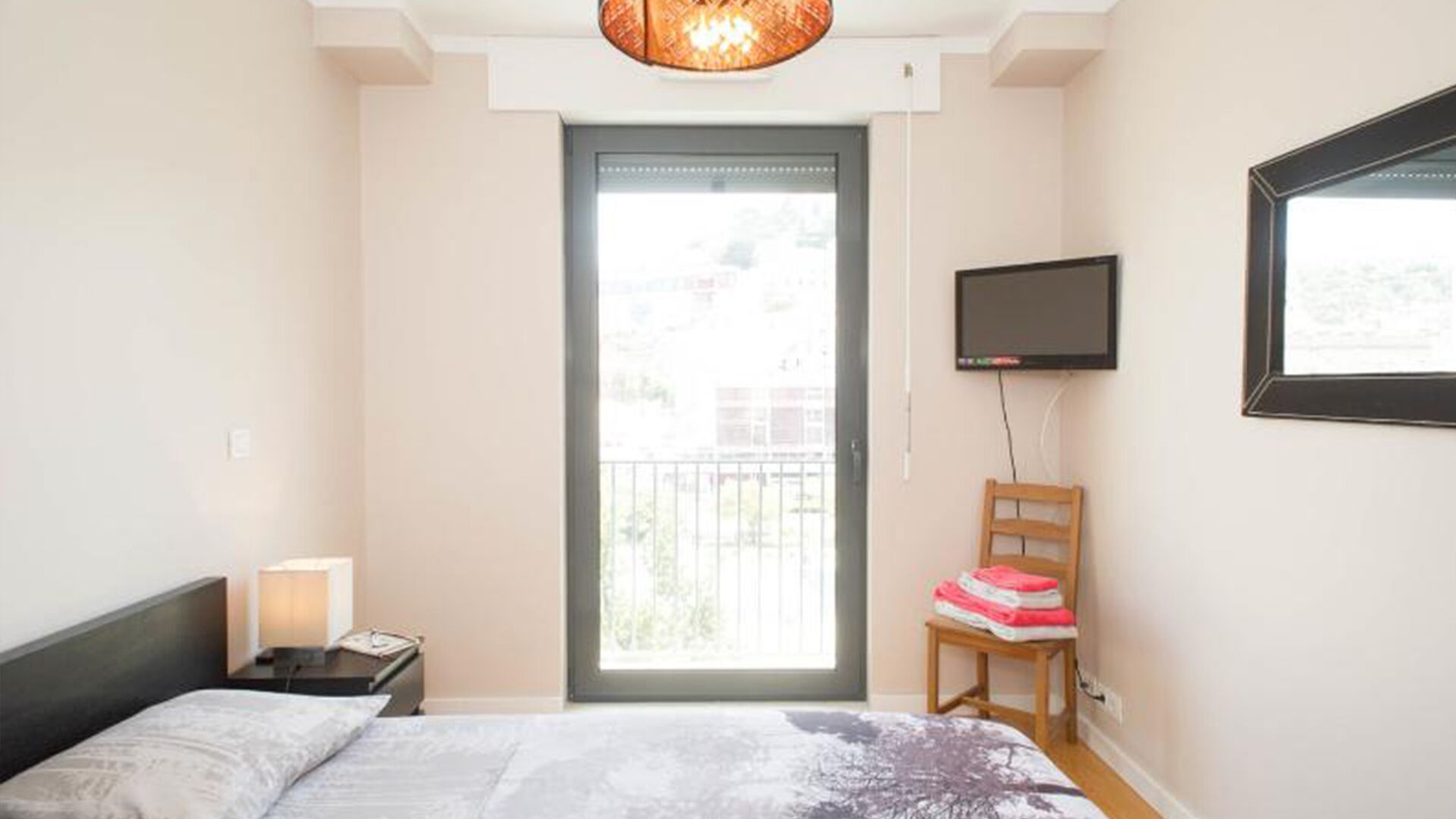 Une chambre à coucher avec un lit, une table de chevet, un téléviseur fixé au mur, une chaise sur laquelle sont pliés des vêtements, et une porte-fenêtre donnant sur un balcon.