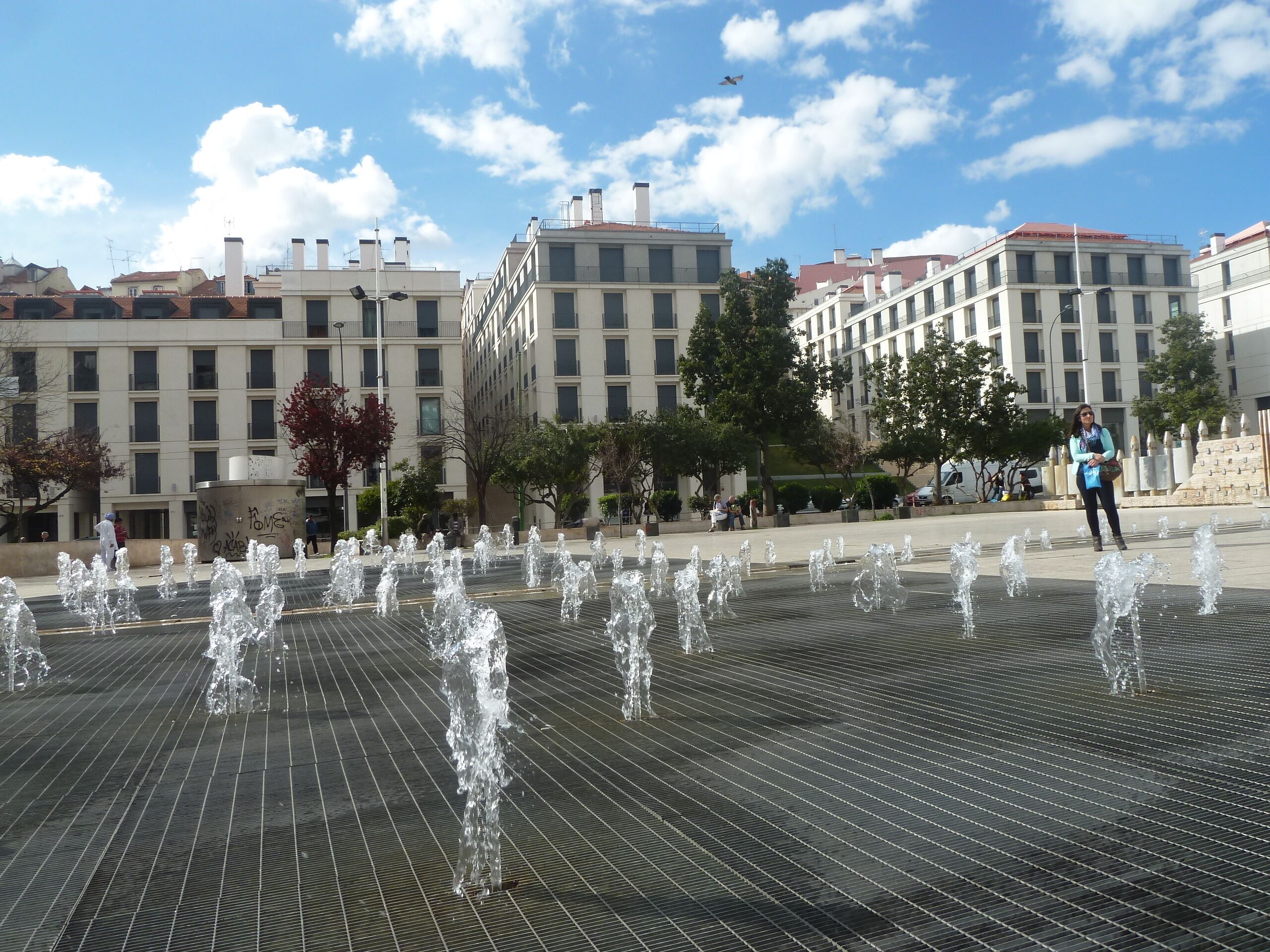 A public square with a water fountain feature, surrounded by modern buildings and trees.