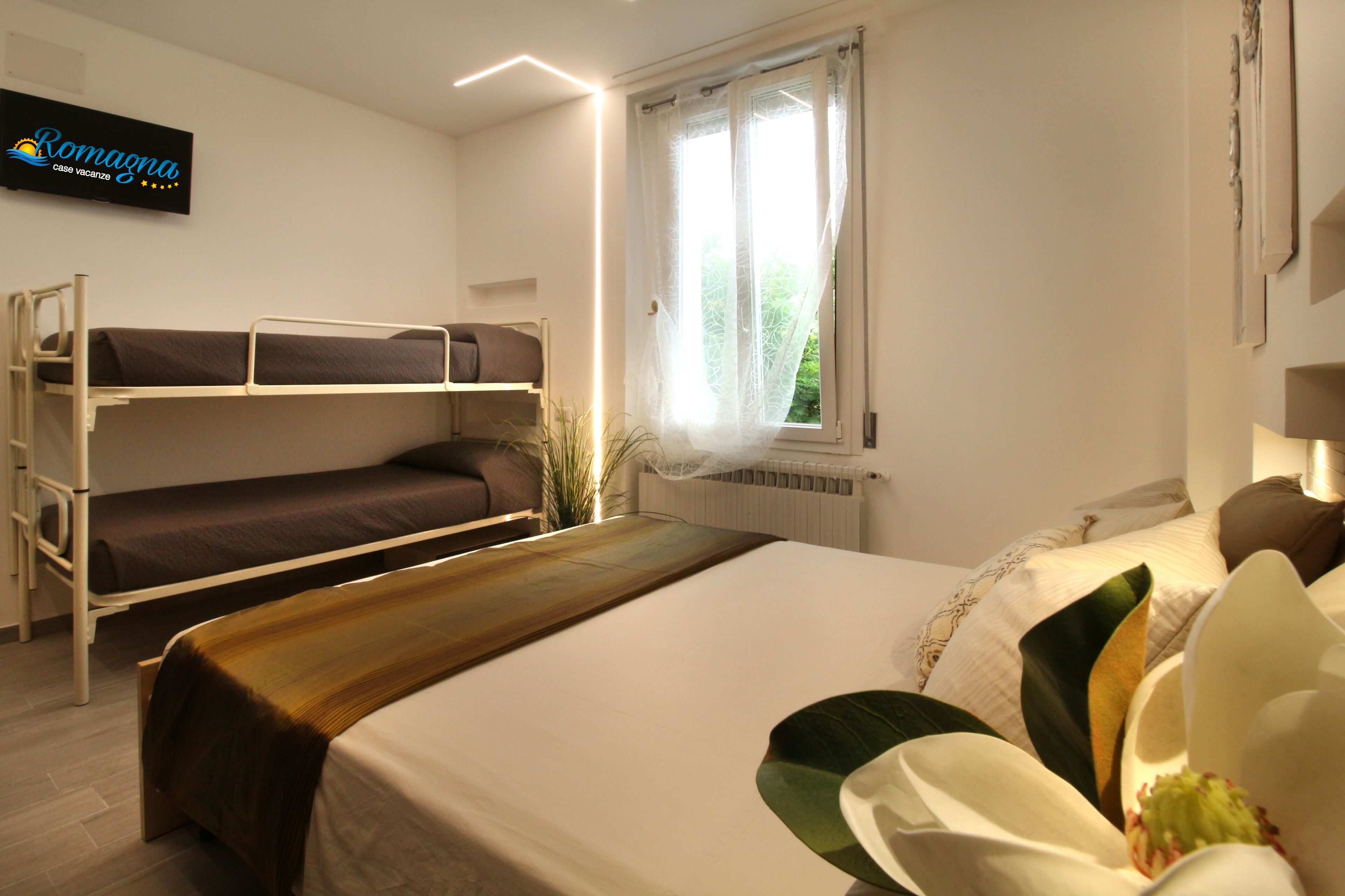 Une chambre d’hôtel avec des lits superposés, un grand lit, une table de chevet en bois, une fenêtre avec des rideaux et un panneau sur le mur.